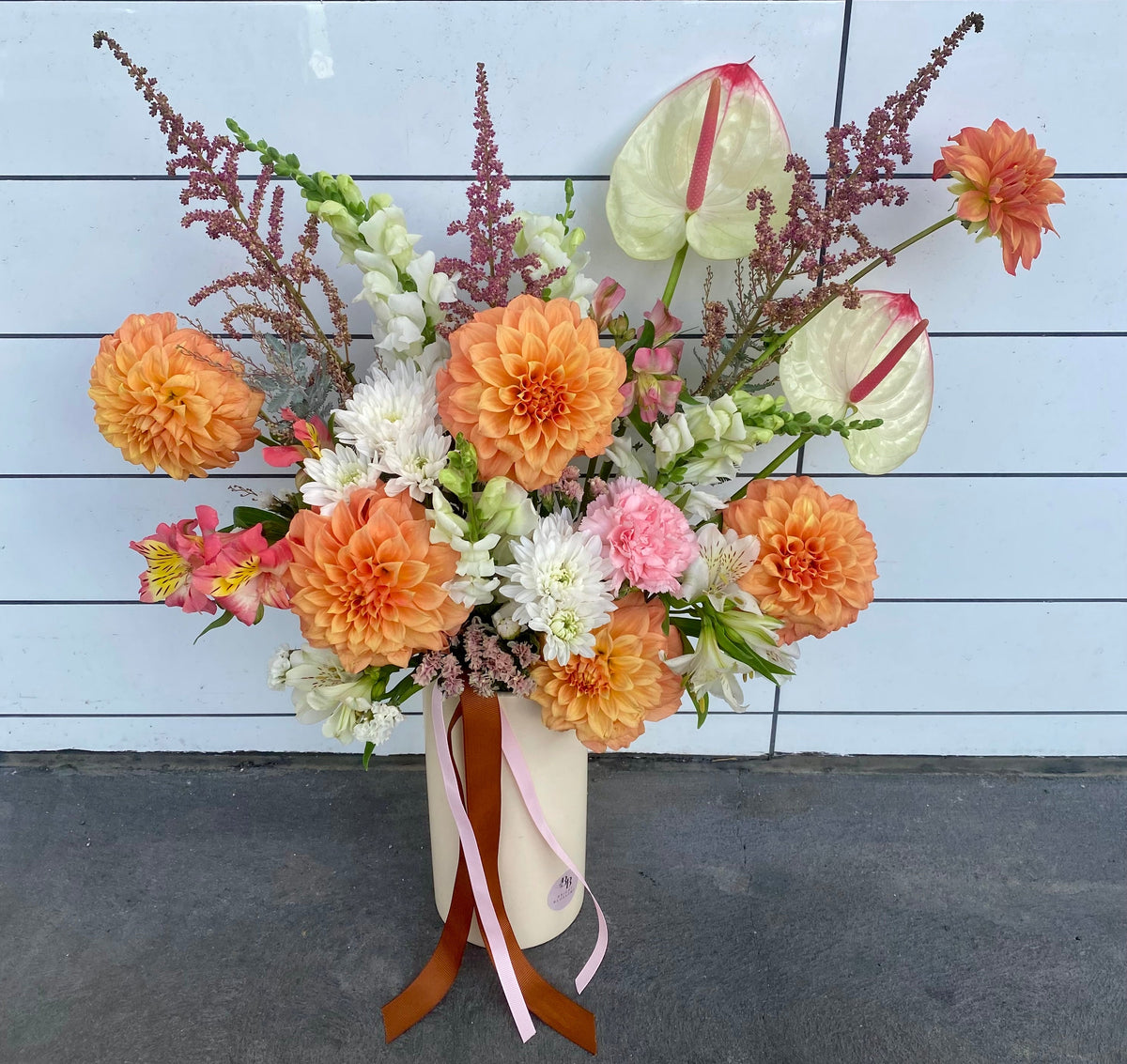 Sunset Delight Vase featuring warm sunset‑toned blooms including dahlias, carnations, chrysanthemums, lilies and anthuriums arranged in a soft cream vase.