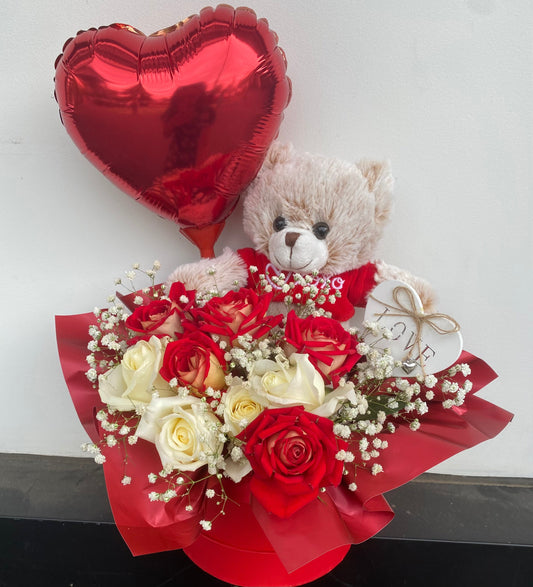 Cuddles & Roses Gift Box featuring a soft plush bear, red heart‑shaped foil balloon, white love‑heart pick and a mix of red and white roses with baby’s breath arranged in an elegant round gift box.
